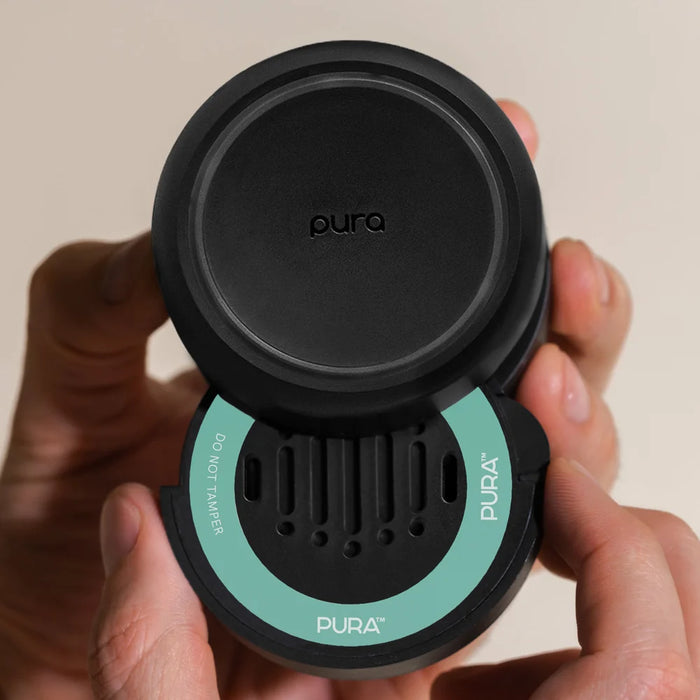 A close-up of a person's hands holding a black circular Pura coffee accessory with a mint green accent ring and the top half lifted off to reveal the interior.