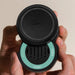 A close-up of a person's hands holding a black circular Pura coffee accessory with a mint green accent ring and the top half lifted off to reveal the interior.