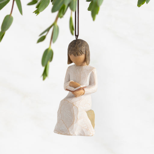 Decorative figurine of a girl holding a book, hanging from a branch with green leaves.