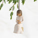 Decorative figurine of a girl holding a book, hanging from a branch with green leaves.