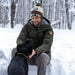 Man sitting in the snow with a dog, wearing a green jacket and a beanie with text.