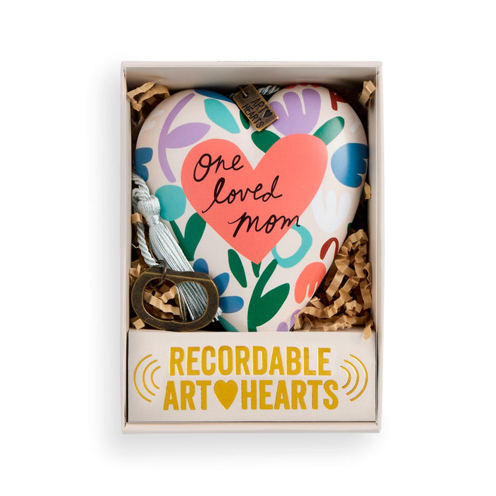 A heart-shaped keepsake ornament presented in a gift box. The ornament has a white base with colorful floral patterns in purple, blue, green, and orange, featuring a large coral-pink heart in the center with the words 'one loved mom' in black script.
