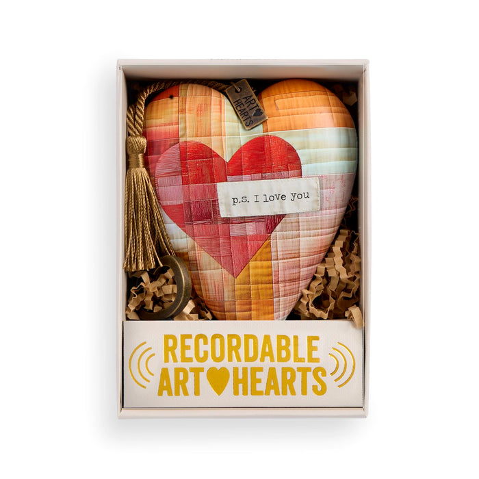 A heart-shaped quilted ornament in a gift box, featuring a patchwork design of red, pink, orange, and yellow plaid sections with a central red heart. The ornament has a hanging loop, a decorative tassel, a small tag, and a paper label that reads 'p.