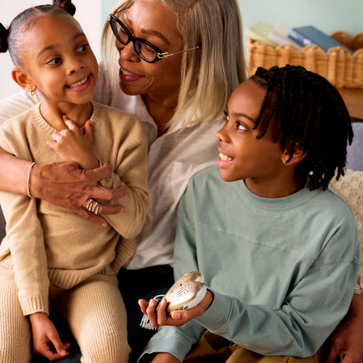A grandmother and her two children are sitting together in a cozy indoor setting. The child on the right is holding a small silver and white decorative object, while the grandmother and the other child look at it with smiles and affection.