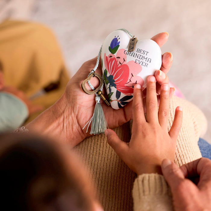 A heart-shaped white keepsake box with floral artwork and the text 'BEST GRANDMA EVER' is held in two hands. The box has a tassel, key ring, and zipper closure, and is shown in a close-up lifestyle setting with a child's hand resting on it.