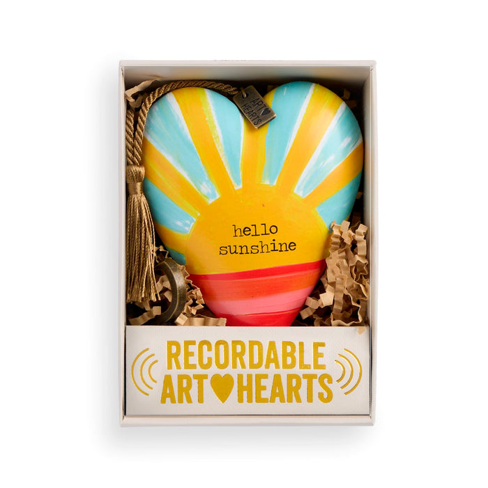 A heart-shaped ceramic ornament with a sunburst design in yellow, light blue, and coral red, displayed inside its white gift box with crinkle paper. The ornament features the text 'hello sunshine' and has a hanging loop with a tassel.