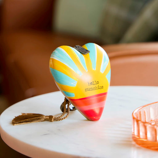 A heart-shaped decorative object with a colorful sunburst pattern in yellow, light blue, and red, sitting on a white marble table. The object features the text 'hello sunshine' in a typewriter font and is styled in a warm, softly blurred indoor setting.