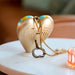 A heart-shaped metallic gold bottle opener pendant with a gold rope chain and a metal keyring, resting on a white marble table.