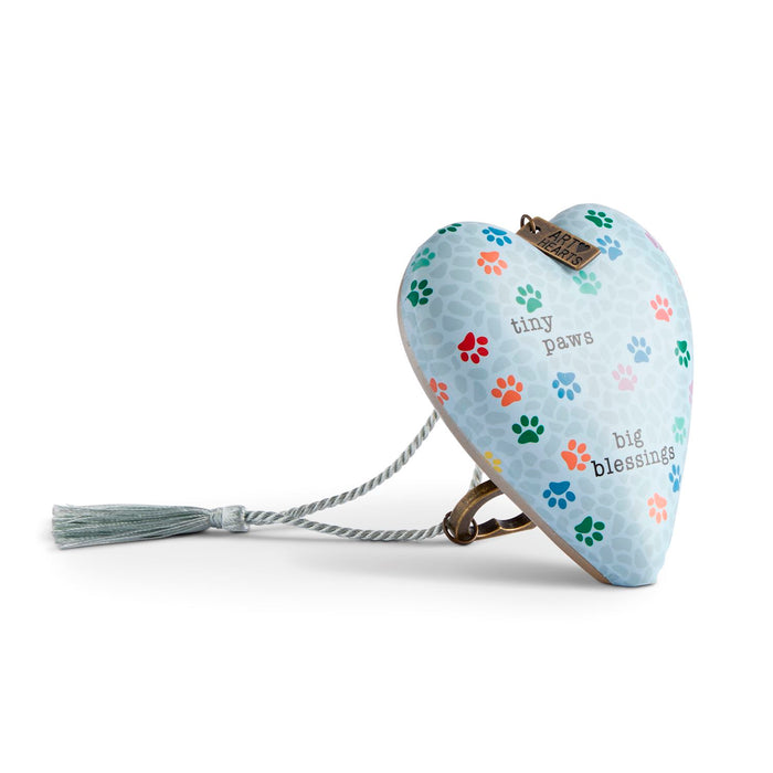 A heart-shaped pet memorial keepsake box with a light blue background, decorated with colorful paw prints and text reading 'tiny paws' and 'big blessings.' It has a gray rope and tassel, attached to a metal clasp, shown on a white background.