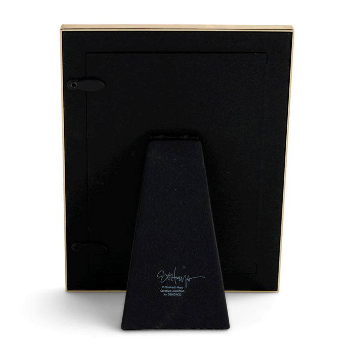 The back view of a black picture frame with a gold-toned border and a matching black triangular stand, featuring a white signature on the stand, set against a plain white background.