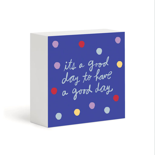 A square decorative block sign with a white side and a blue front panel. The front features the phrase 'its a good day to have a good day' in white script, surrounded by scattered pastel and bright polka dots in red, yellow, light blue, and lavender.