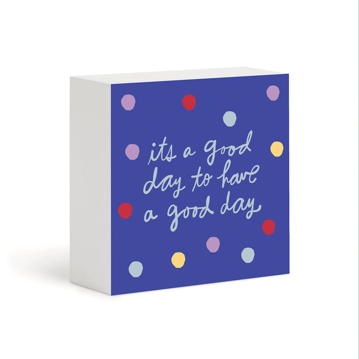 A square decorative block sign with a white side and a blue front panel. The front features the phrase 'its a good day to have a good day' in white script, surrounded by scattered pastel and bright polka dots in red, yellow, light blue, and lavender.