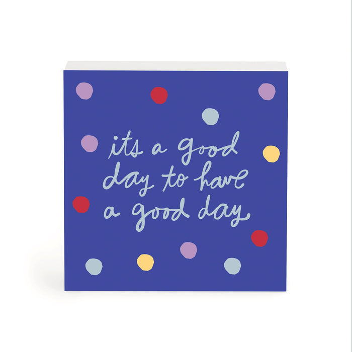 A square blue sign or plaque with the phrase “it’s a good day to have a good day” in white script, decorated with scattered pastel and bright polka dots in purple, red, yellow, and light blue, shown on a white background.