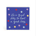 A square blue sign or plaque with the phrase “it’s a good day to have a good day” in white script, decorated with scattered pastel and bright polka dots in purple, red, yellow, and light blue, shown on a white background.