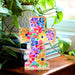 Colorful floral cross decoration on a wooden surface with books and plants in the background