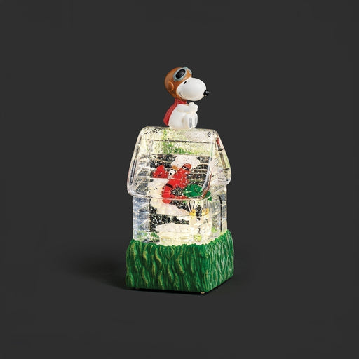A Snoopy doghouse snow globe figurine featuring Snoopy in a brown aviator hat and red scarf perched atop a clear glass house with a green grass-textured base, set against a dark gray background.