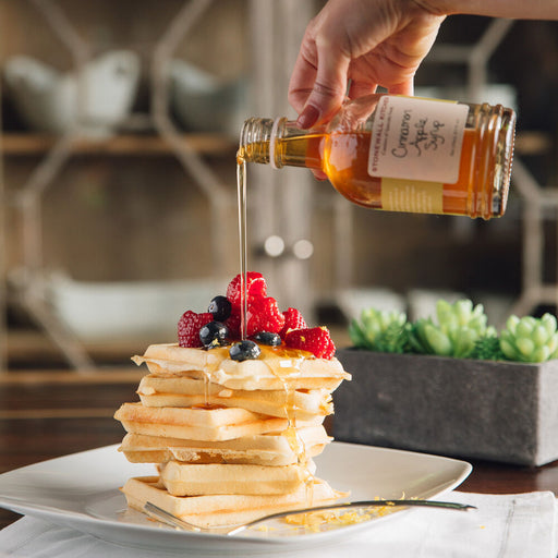 Person pouring syrup over a stack of pancakes with berries on a plate.