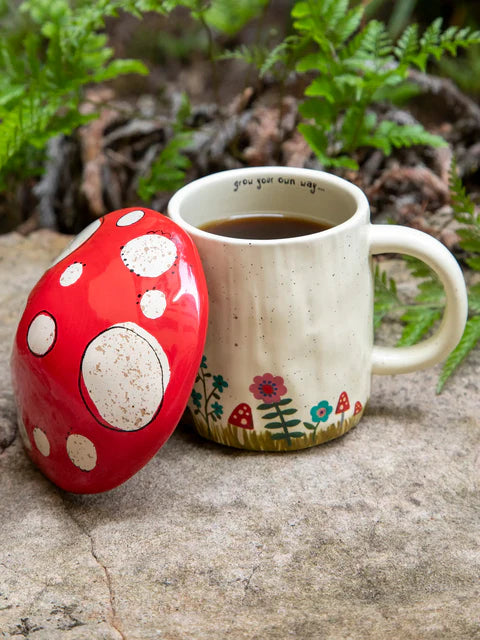 Red mushroom-shaped container and ceramic mug with floral design on a stone surface with greenery in the background