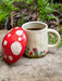 Red mushroom-shaped container and ceramic mug with floral design on a stone surface with greenery in the background