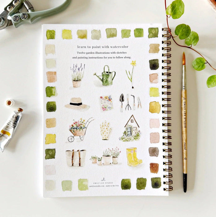 Notebook with watercolor illustrations of garden-themed designs on a white surface with art supplies.