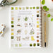 Notebook with watercolor illustrations of garden-themed designs on a white surface with art supplies.