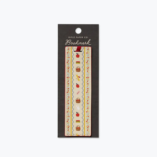 A decorative bookmark from Rifle Paper Co. in its original black hanging card packaging, featuring a vertical strip with a cream background and a whimsical pattern of red apples, brown baskets, and small yellow flowers.