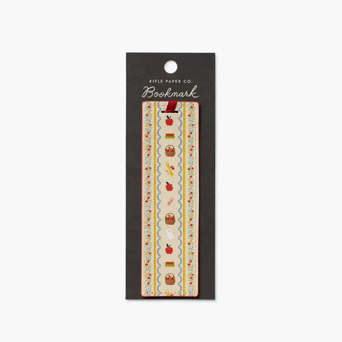 A decorative bookmark from Rifle Paper Co. in its original black hanging card packaging, featuring a vertical strip with a cream background and a whimsical pattern of red apples, brown baskets, and small yellow flowers.