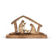 A wooden nativity scene figurine featuring Mary, Joseph, and baby Jesus under a stable roof silhouette, crafted from natural wood with a rustic finish, set against a plain white background.