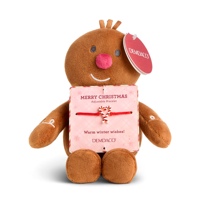 A brown plush gingerbread person toy holding a pink Christmas gift box with a red candy cane bow. The toy has a round pink nose, embroidered white details on its arms, and a red hanging tag that reads DEMDACO.