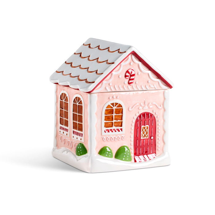 A gingerbread house ceramic container or ornament in light pink and white, featuring a red front door, arched windows with brown trim, green bushes, and a candy cane detail on the roof.