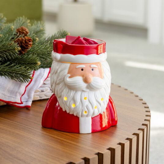 A Santa Claus-shaped ceramic candle holder sits on a wooden table, with green pine branches and a white cloth nearby in a softly blurred indoor setting.