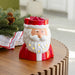 A Santa Claus-shaped ceramic candle holder sits on a wooden table, with green pine branches and a white cloth nearby in a softly blurred indoor setting.
