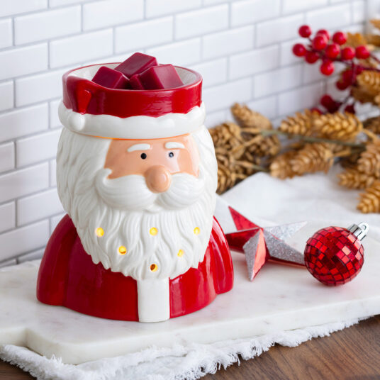 A Santa Claus-shaped ceramic candle holder with built-in warm lights, styled on a marble board with Christmas decorations in the background.