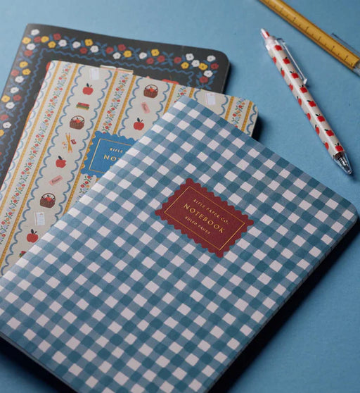 A stack of patterned notebooks on a blue surface, with the top notebook in blue and white gingham featuring a red label that reads 'MITE RIKO CO. NOTEPAD'.