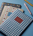 A stack of patterned notebooks on a blue surface, with the top notebook in blue and white gingham featuring a red label that reads 'MITE RIKO CO. NOTEPAD'.