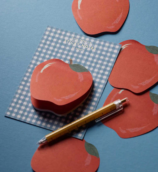 A styled flat-lay of red apple-shaped sticky notes on a blue background, with one note on a blue-and-white gingham sheet and a gold pen placed diagonally across the scene.
