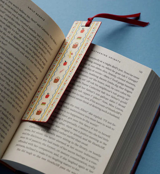 A decorative paper bookmark with a red ribbon loop, featuring a whimsical pattern of red apples and brown bears on a yellow and blue wavy border, resting on open book pages against a solid blue background.