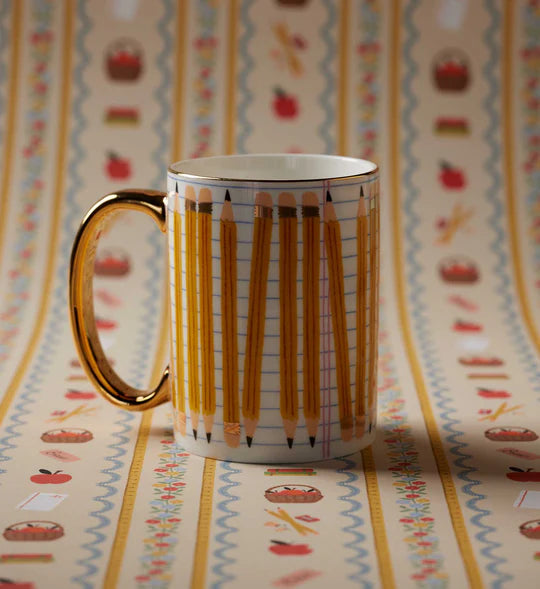 A white ceramic mug with a gold handle and gold rim, decorated with a pattern of yellow pencils on graph paper, set against a matching patterned background.