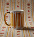 A white ceramic mug with a gold handle and gold rim, decorated with a pattern of yellow pencils on graph paper, set against a matching patterned background.