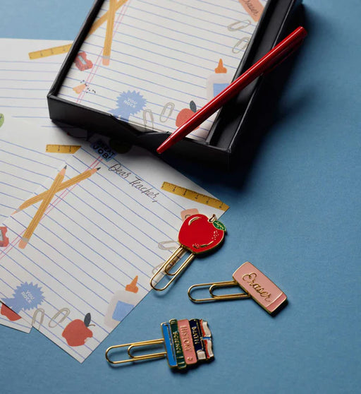 A styled flat-lay of school-themed paperclip bookmarks and a red pen on patterned notepad sheets over a blue background.