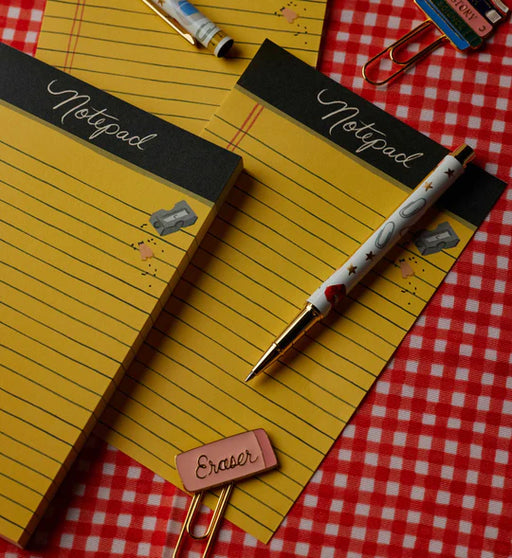 A styled flat-lay of yellow lined notepad pages with black headers reading 'Notepad' and decorative doodles, accompanied by a white pen with gold trim, paper clips, and a small pink eraser on a red-and-white gingham surface.