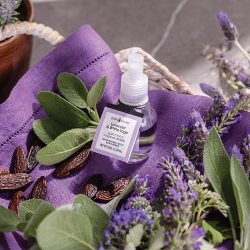 Bottle of lavender and white sage essential oil spray on a purple cloth with lavender flowers and leaves.