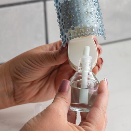 Hand holding a small glass bottle with a white cap, attached to a decorative blue light fixture.