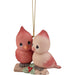 A Christmas ornament featuring two cute, stylized birds perched on a small branch with red berries, hanging from a gold string against a plain white background.