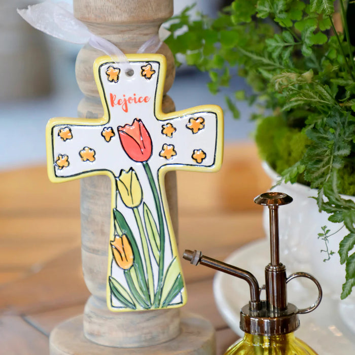 Decorative cross with floral design and 'Rejoice' text on a wooden stand with a plant in the background.