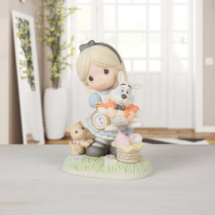 A Precious Moments figurine of a blonde girl in a blue dress holding a white rabbit, with a small bear and a basket on a grassy base, displayed on a tabletop in a softly lit room.