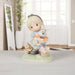 A Precious Moments figurine of a blonde girl in a blue dress holding a white rabbit, with a small bear and a basket on a grassy base, displayed on a tabletop in a softly lit room.