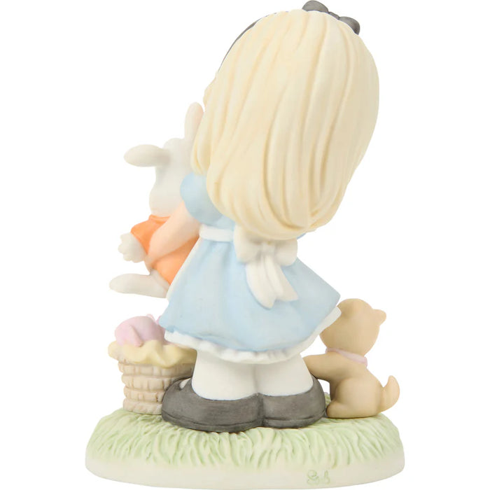 A small figurine of a young girl with blonde hair seen from behind, wearing a light blue dress and gray shoes, standing on a grassy base with a basket, a white rabbit, and a teddy bear.