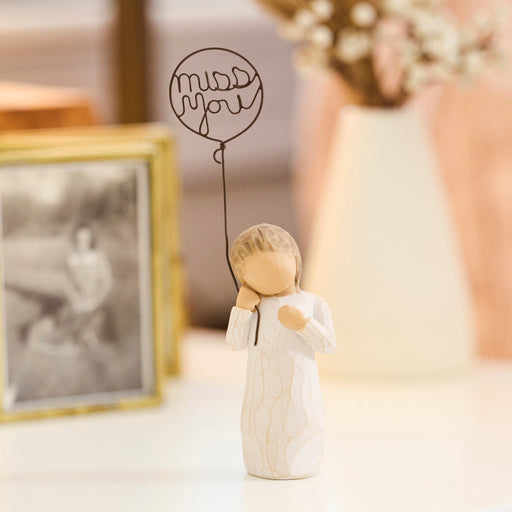 Small figurine holding a balloon with 'miss you' text, next to a framed photo on a table.