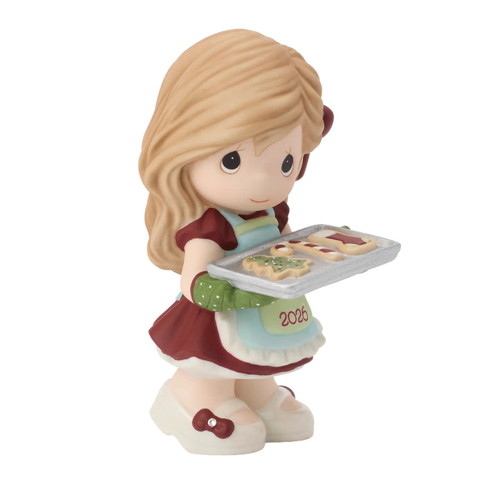 A Precious Moments figurine of a young girl with blonde hair holding a tray of cookies, wearing a red dress, green gloves, and a light blue apron with '2006' written on it, set against a plain white background.